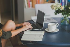 Female freelancer using laptop with coffee at home office desk, surrounded by roses and a planner.